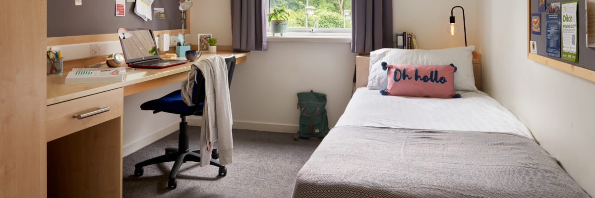 An empty student bedroom has a bed on the right-hand side, a desk and bookshelves on the left.