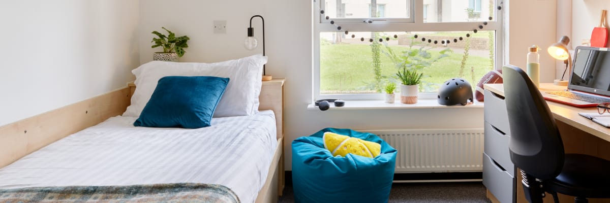 An empty student bedroom has a bed on the left-hand side, a desk and bookshelves on the right.
