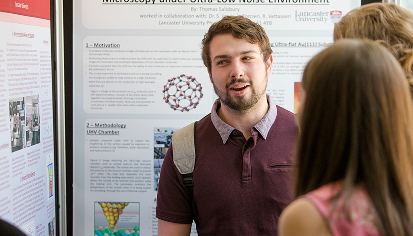 A male student stands in front of his poster, talking to fellow students