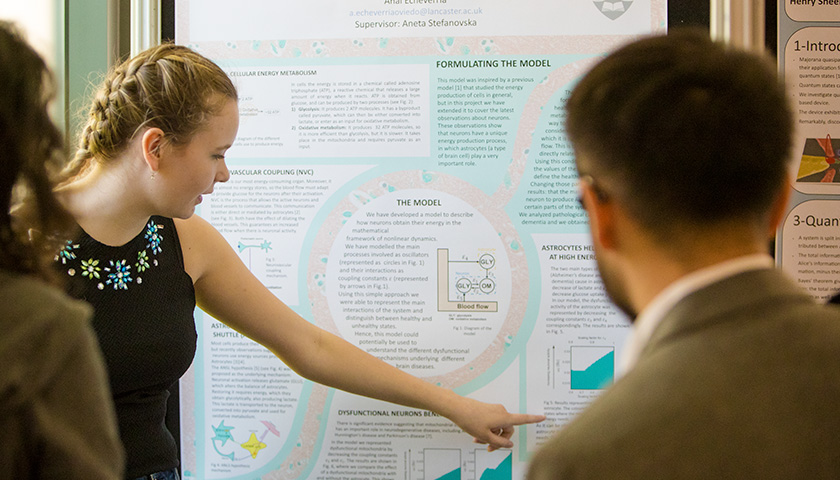 A female student pointing at her poster whilst a lecturer looks on