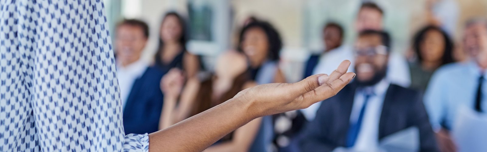 A person gesticulating with their right hand in front of an out of focus audience of businesspeople
