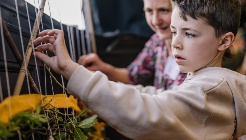 a child weaves some twigs between string, adding to a wall hanging display