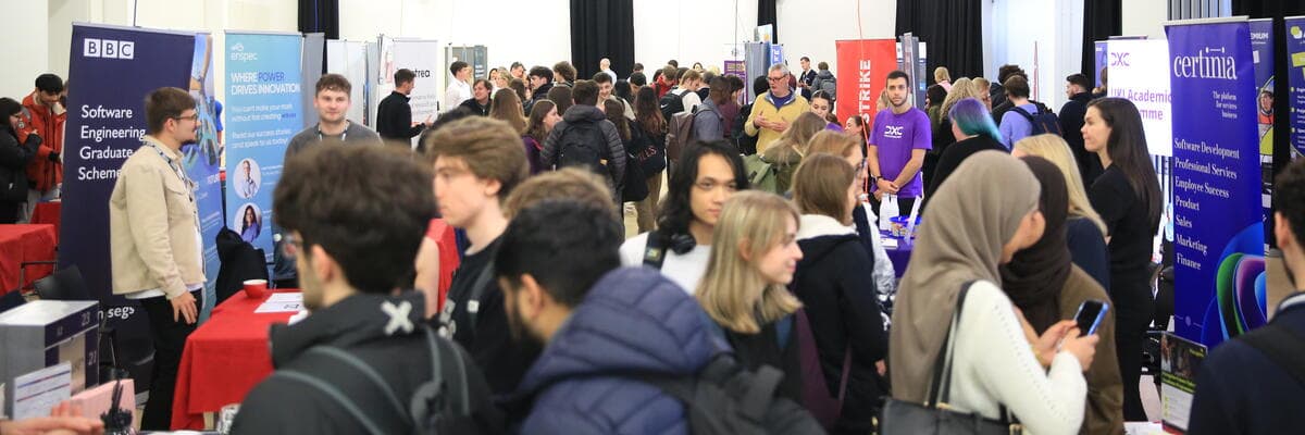 Students speak with an exhibitor at an indoor careers fair, standing beside recruitment display banners in a busy event space.
