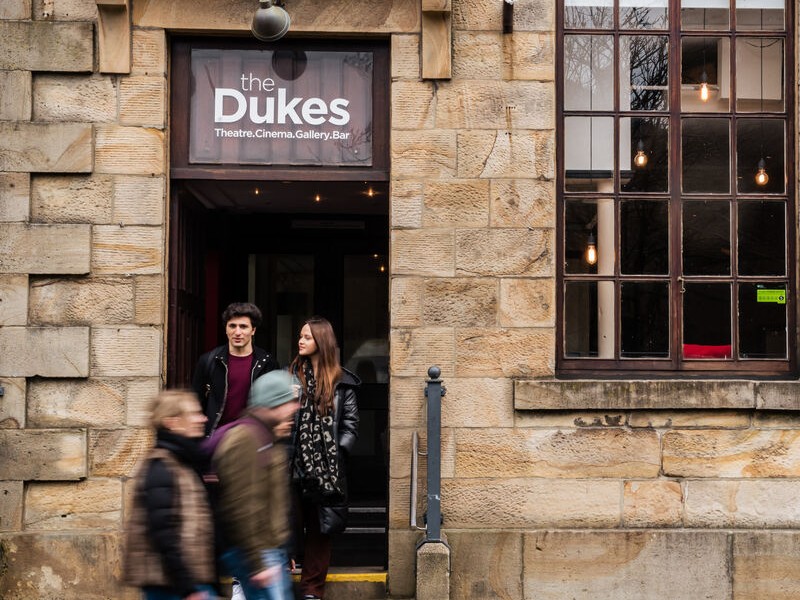 Two people at the entrence of the Dukes theatre and two people walking pass in front of them.