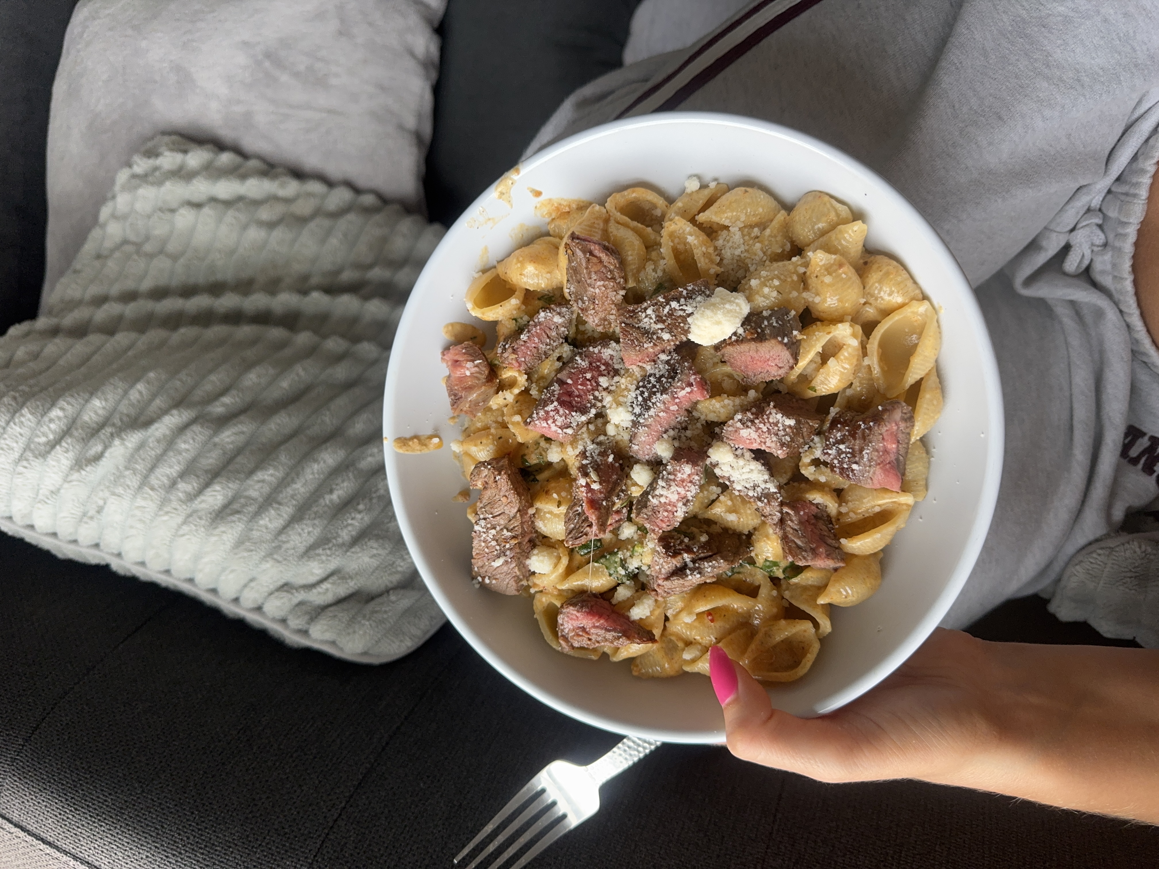 A person holding a bowl of steak pasta sprinkled with cheese on a cozy couch.
