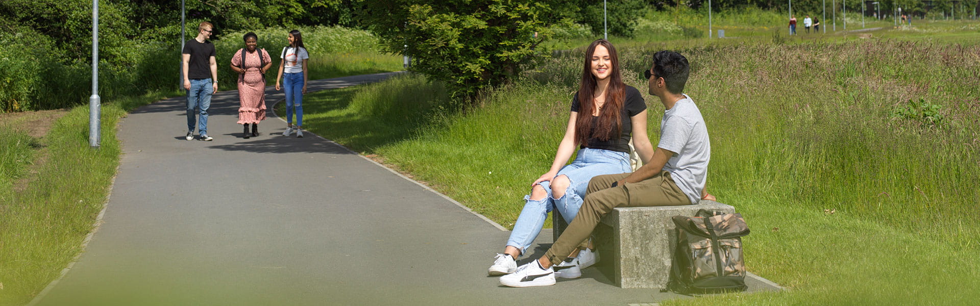 Students chat on a bench in the sunshine surrounded by landscaped parkland