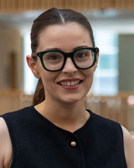 Head shot of Ophelia Chidgey, she has tied back black hair, thick framed glasses, she is smiling.