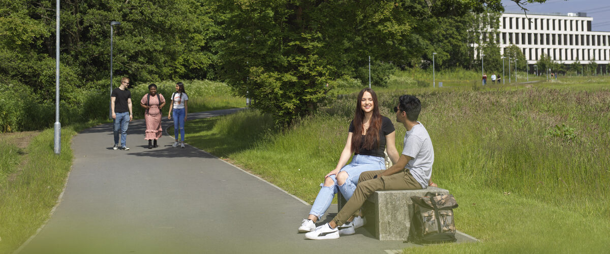 Students sit on a bench in the campus parkland. More people walk along a path in the background.